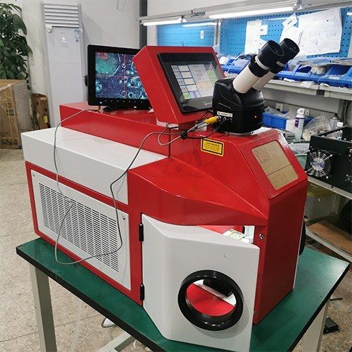 Desktop jewelry laser welder on a workshop bench with a second monitor showing a magnified view of the part